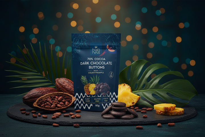 A 80g pouch of Nova Nova 70% Cocoa Dark Chocolate Buttons with Golden Pineapple flavor, displayed on a wooden table next to several dark chocolate buttons.